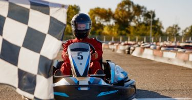A person wearing a black helmet sits in a go-kart on an outdoor track behind a black and white checkered flag
