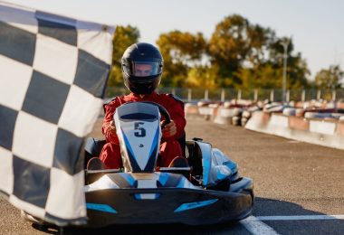 A person wearing a black helmet sits in a go-kart on an outdoor track behind a black and white checkered flag