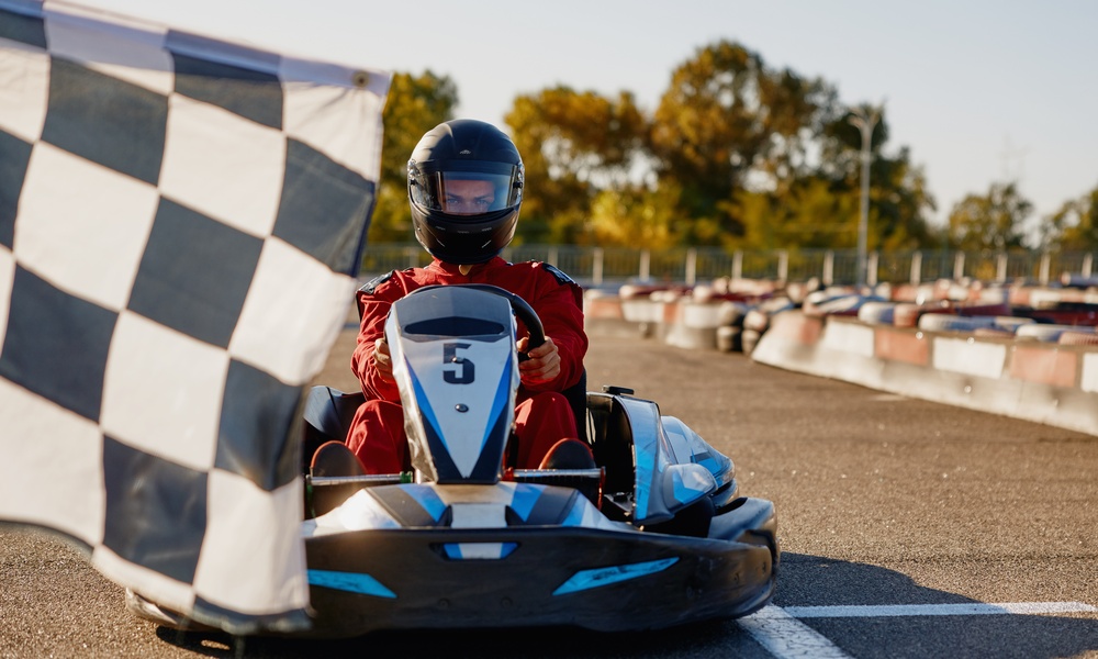 A person wearing a black helmet sits in a go-kart on an outdoor track behind a black and white checkered flag