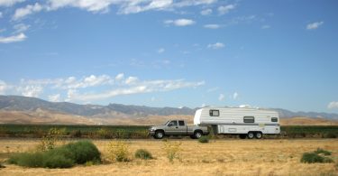 A silver pickup truck towing a white motorhome down a rural desert highway under a bright blue sky with a few clouds.