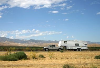 A silver pickup truck towing a white motorhome down a rural desert highway under a bright blue sky with a few clouds.