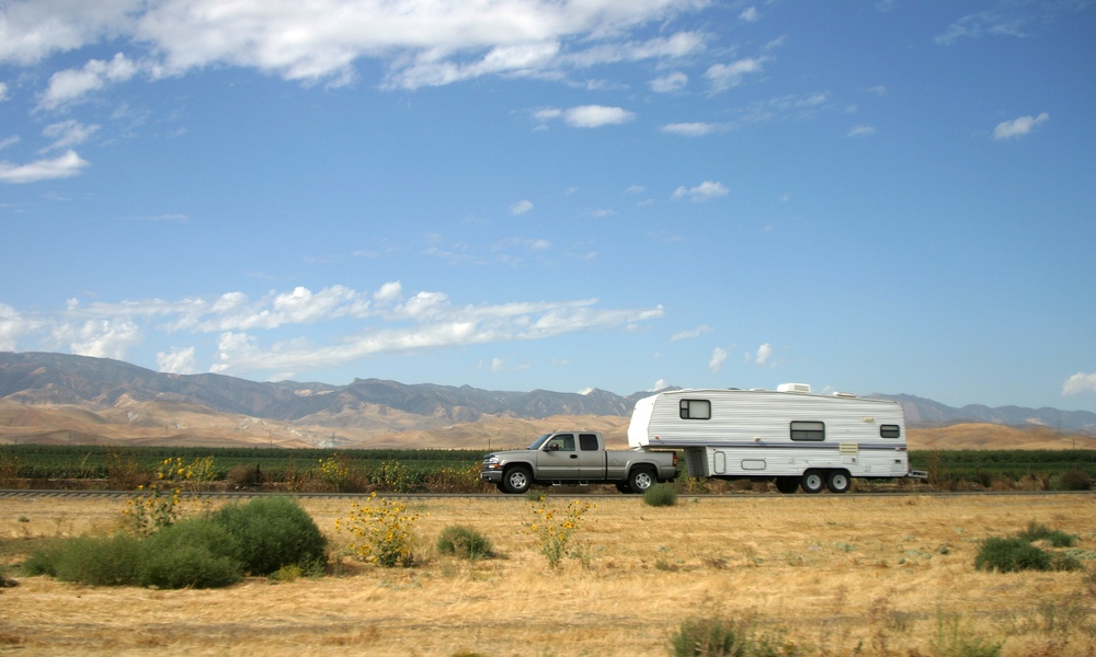 A silver pickup truck towing a white motorhome down a rural desert highway under a bright blue sky with a few clouds.