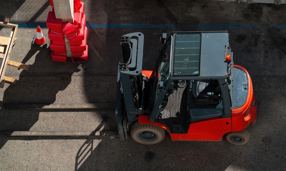 A red forklift moves pallets in an outdoor industrial area. Safety barriers and concrete ground are visible.