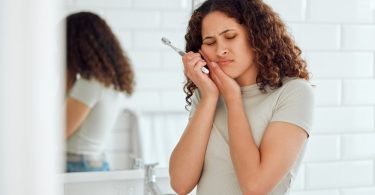 A woman brushing her teeth while holding her cheek in a bathroom, with dental hygiene items on the counter.