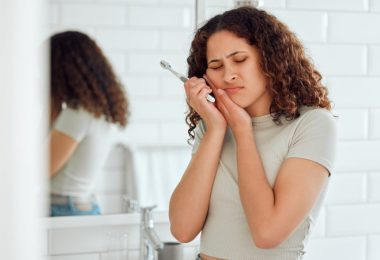 A woman brushing her teeth while holding her cheek in a bathroom, with dental hygiene items on the counter.