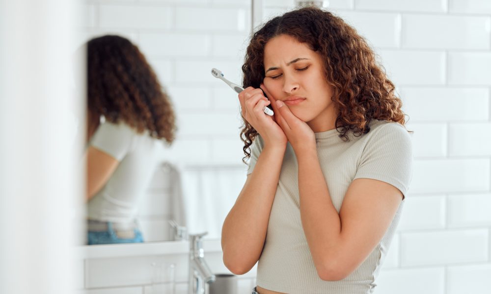 A woman brushing her teeth while holding her cheek in a bathroom, with dental hygiene items on the counter.