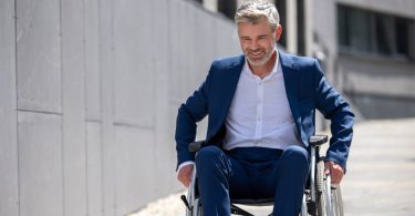 A well-dressed man in a wheelchair, wheeling himself down an outdoor ramp. He has both hands on the wheels.