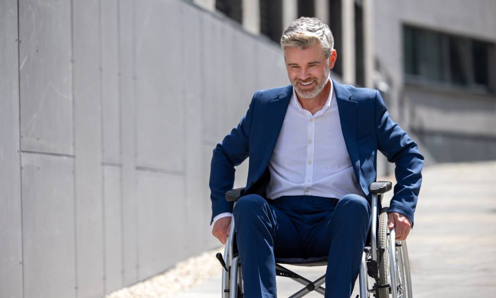 A well-dressed man in a wheelchair, wheeling himself down an outdoor ramp. He has both hands on the wheels.