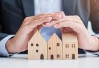 A man in a suit using his hands to create a cover over three small wooden blocks shaped like houses below him.