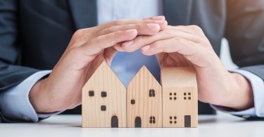 A man in a suit using his hands to create a cover over three small wooden blocks shaped like houses below him.