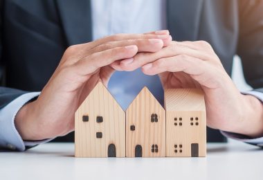 A man in a suit using his hands to create a cover over three small wooden blocks shaped like houses below him.
