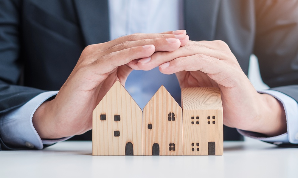 A man in a suit using his hands to create a cover over three small wooden blocks shaped like houses below him.