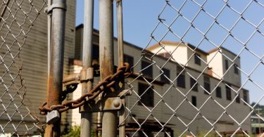 a rusted chain and padlock securing a metal fence in front of an old industrial building under a blue sky.
