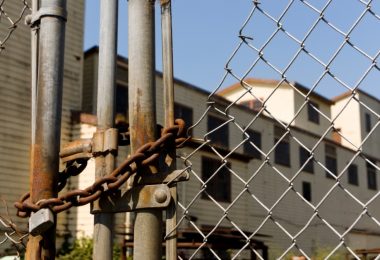 a rusted chain and padlock securing a metal fence in front of an old industrial building under a blue sky.