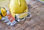 A collection of PPE gear on a wooden work surface, such as a yellow hard hat, safety glasses, and gloves.