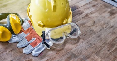 A collection of PPE gear on a wooden work surface, such as a yellow hard hat, safety glasses, and gloves.