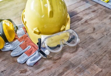 A collection of PPE gear on a wooden work surface, such as a yellow hard hat, safety glasses, and gloves.