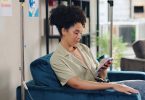 a woman sitting in a blue chair beside an IV drip while looking at a smartphone, with a sofa in the background.