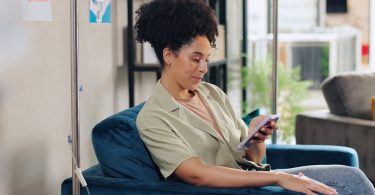 a woman sitting in a blue chair beside an IV drip while looking at a smartphone, with a sofa in the background.