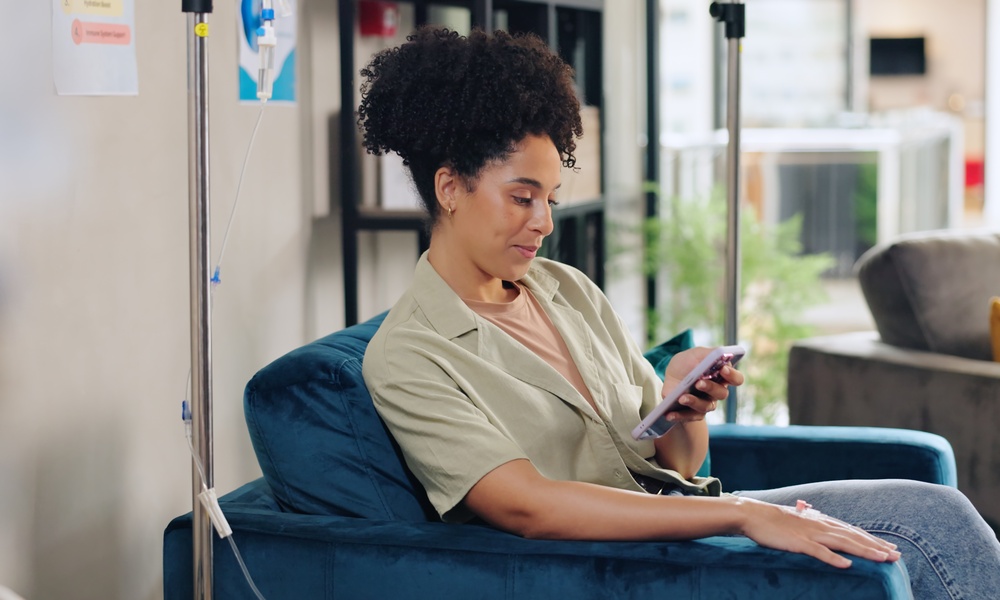 a woman sitting in a blue chair beside an IV drip while looking at a smartphone, with a sofa in the background.
