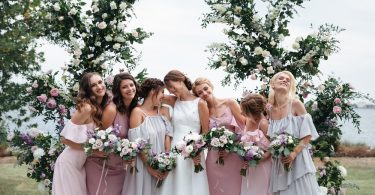 A group of seven women in pastel dresses holding flower bouquets in front of tall floral arrangements.