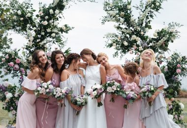 A group of seven women in pastel dresses holding flower bouquets in front of tall floral arrangements.