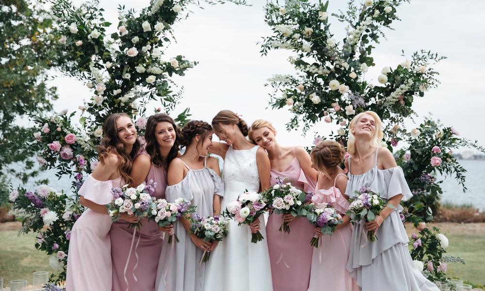 A group of seven women in pastel dresses holding flower bouquets in front of tall floral arrangements.