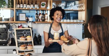 In a cafe, a barista is serving a customer her order. There is a small display with muffins on the counter.