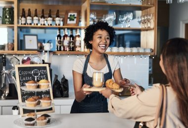In a cafe, a barista is serving a customer her order. There is a small display with muffins on the counter.
