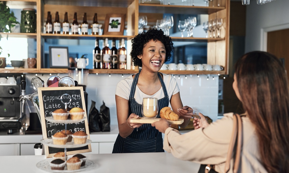 In a cafe, a barista is serving a customer her order. There is a small display with muffins on the counter.
