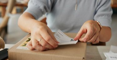 A person is placing a tracking sticker on a cardboard box. There are papers to the side of the box on a table.