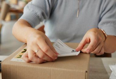 A person is placing a tracking sticker on a cardboard box. There are papers to the side of the box on a table.
