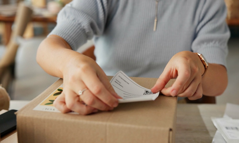 A person is placing a tracking sticker on a cardboard box. There are papers to the side of the box on a table.