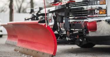 a red snowplow blade attached to a pickup truck parked on pavement with a second truck blurred behind.