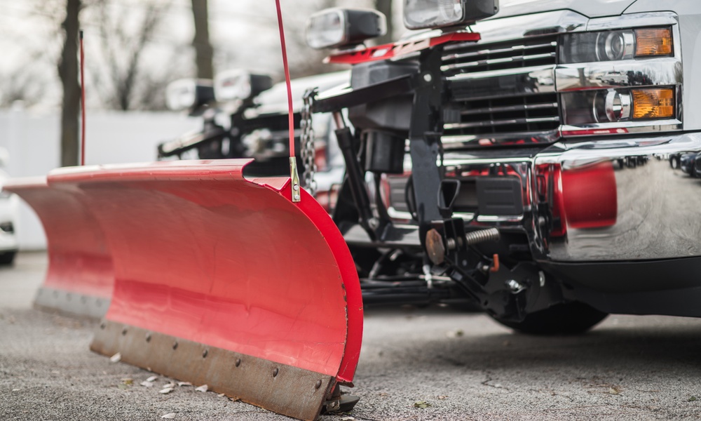 a red snowplow blade attached to a pickup truck parked on pavement with a second truck blurred behind.