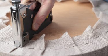 A hand pressing a metal staple gun into beige fabric wrapped around a round wooden seat during upholstery work.