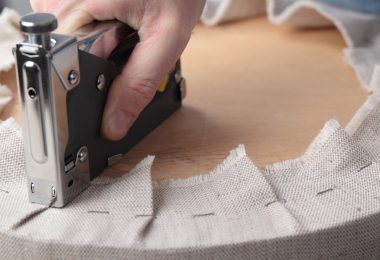 A hand pressing a metal staple gun into beige fabric wrapped around a round wooden seat during upholstery work.
