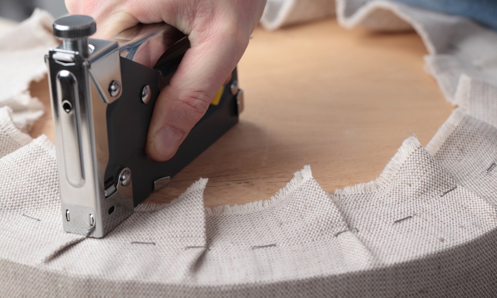 A hand pressing a metal staple gun into beige fabric wrapped around a round wooden seat during upholstery work.