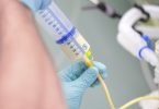 Someone wearing blue latex gloves connects a syringe of pale yellow liquid to a feeding tube.