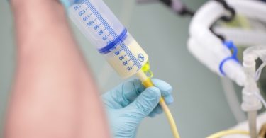 Someone wearing blue latex gloves connects a syringe of pale yellow liquid to a feeding tube.