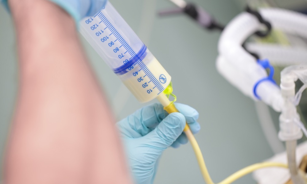 Someone wearing blue latex gloves connects a syringe of pale yellow liquid to a feeding tube.