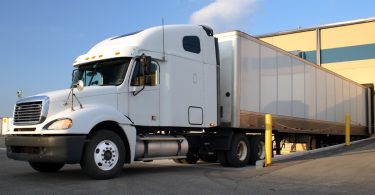 A white semi truck with a box trailer parked beside a warehouse loading dock under a blue sky with clouds.