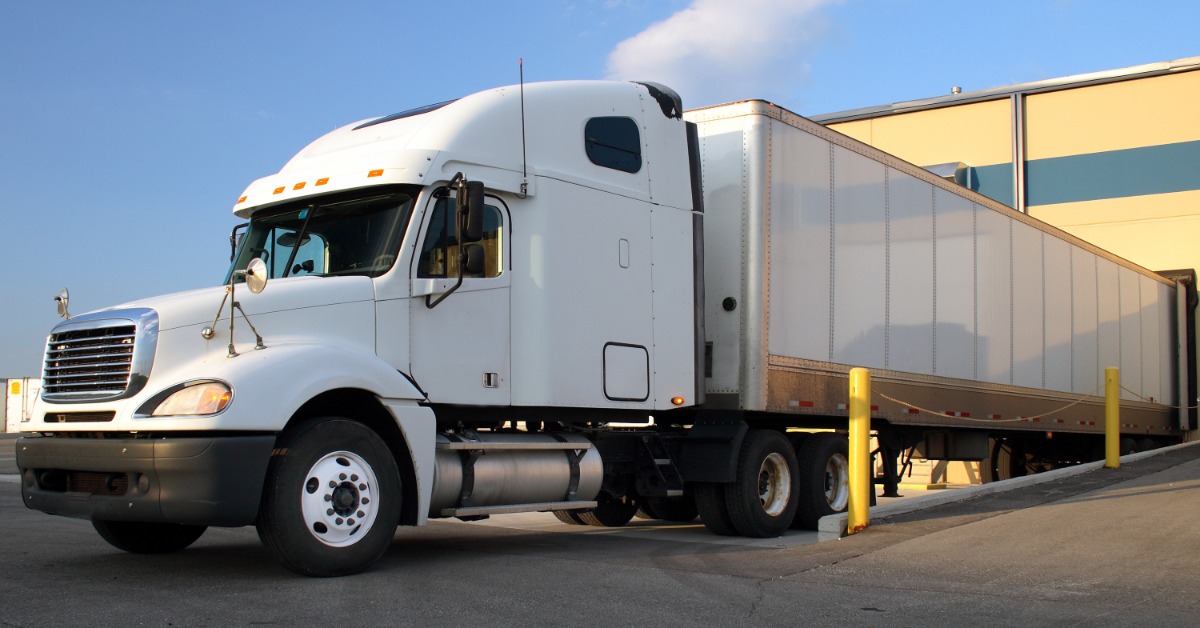 A white semi truck with a box trailer parked beside a warehouse loading dock under a blue sky with clouds.