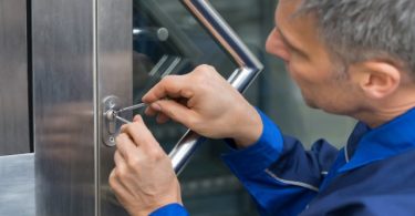 A close-up of a man wearing blue coveralls while using lock picking tools to work on a lock on a metal-framed door.