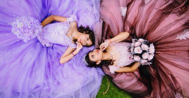Two young women in elegant quinceañera gowns lie on green grass, with flowing purple and rose skirts spread in circles.