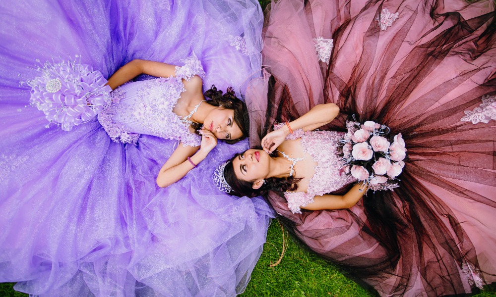 Two young women in elegant quinceañera gowns lie on green grass, with flowing purple and rose skirts spread in circles.