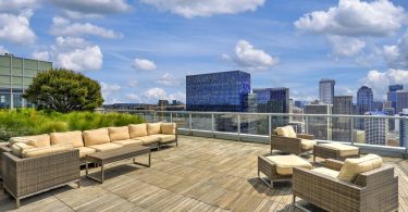 Rooftop apartment patio with cushioned lounge seating, wood decking, glass railing, and a city skyline under blue skies.