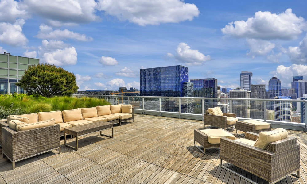 Rooftop apartment patio with cushioned lounge seating, wood decking, glass railing, and a city skyline under blue skies.