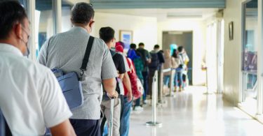 Travelers stand in a crowded airport terminal line with backpacks, luggage, seats, and digital signs nearby.