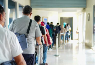 Travelers stand in a crowded airport terminal line with backpacks, luggage, seats, and digital signs nearby.